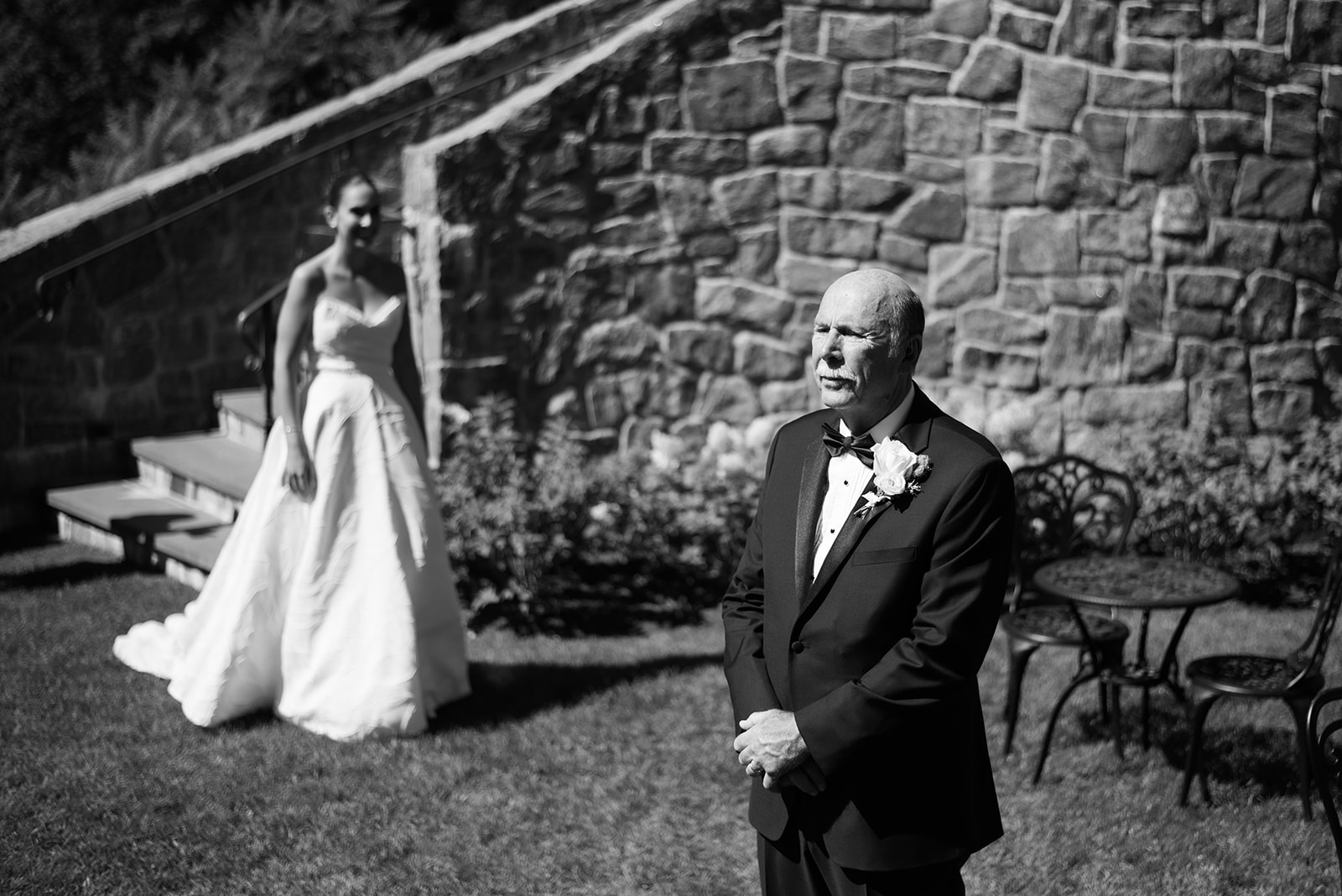 Black and white portrait of a father's emotional first look as the bride stands on garden stairs behind him.